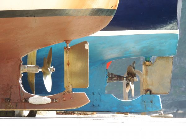 Fotografía artística de hélices de barco vistas desde abajo, con contrastes de colores azul y óxido, enmarcada sobre fondo blanco.